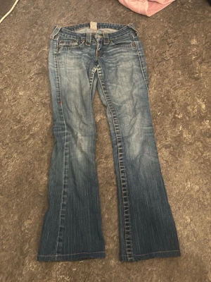 Blå bootcut jeans från true religion - Säljer ett par blå bootcut jeans från true religion med slitningar och ljusare partier framtill. Klassisk femficksmodell med låg midja och utsvängda ben. Jeansen har detaljerade sömmar och är tillverkade i denim med en skön känsla.