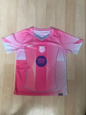 FC Barcelona rosa matchtröja Nike - Snygg FC Barcelona matchtröja i rosa och vitt med gradientmönster och tunna linjer. Tröjan har klubbmärke, Nike-logga och Spotify-logga på bröstet. Tillverkad i lätt och ventilerande funktionsmaterial, perfekt för träning eller match.