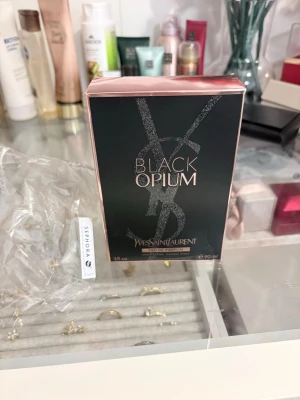 Yves Saint Laurent Black Opium EdP - Yves Saint Laurent Black Opium Eau de Parfum i en elegant svart och roséguldig förpackning. Doften är känd för sina intensiva och sensuella toner med inslag av kaffe, vanilj och vita blommor. Flaskan rymmer 90 ml och har en lyxig design med glittrande detaljer.