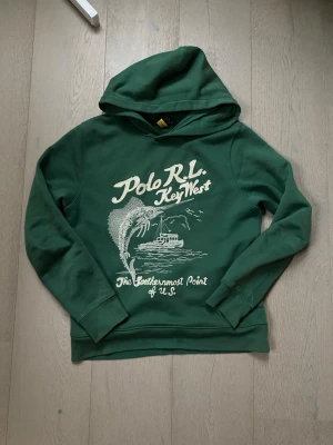 Grön hoodie Polo Ralph Lauren - Snygg grön hoodie från Polo Ralph Lauren med tryck framtill som visar en fisk, båt och texten 'Key West'. Hoodien har huva och ribbade muddar vid ärmar och nederkant. Perfekt för dig som gillar streetwear och vill sticka ut med ett coolt motiv. Känns som storlek xs/s