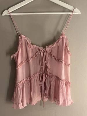 Topp från Zara - Supersöt ljusrosa topp med tunna axelband och volangdetaljer. Toppen har dekorativa sömmar med små volanger och knytning framtill. Tillverkad i ett tunt, luftigt material som känns som polyester eller syntet. Perfekt för varma dagar eller sommarens alla tillfällen.