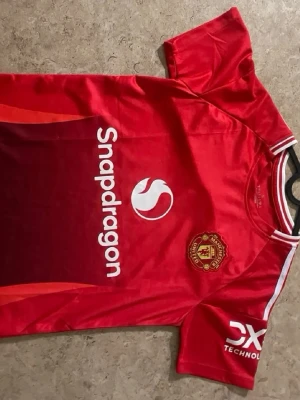 Manchester United röd matchtröja - Röd Manchester United matchtröja med Snapdragon-logga på framsidan och klubbemblem på bröstet. Tröjan har vita detaljer på ärmarna och är tillverkad i ett lätt och ventilerande syntetmaterial. Perfekt för fotbollsträning eller att bära på läktaren.