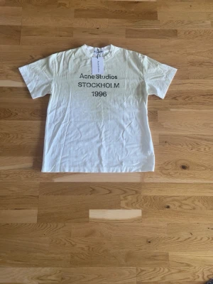 Acne Studios t-shirt - Vit t-shirt från Acne Studios med trycket 'Acne Studios STOCKHOLM 1996' på bröstet. Klassisk passform med rund hals och korta ärmar. Tillverkad i mjuk bomull, perfekt för en clean och stilren look.