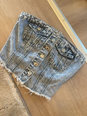Jeans tubtopp med knappar och rå kant - Säljer en snygg tubtopp i ljusblå denim med knappar framtill, två bröstfickor och rå, fransig nederkant. Toppen har smockad rygg för skön passform och cool vintagekänsla. Perfekt att styla till höga jeans eller kjol.