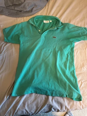 Turkos pikétröja från Lacoste - Snygg turkos pikétröja från Lacoste med klassisk krage och knappar framtill. Ikoniska krokodilloggan broderad på bröstet. Tröjan har korta ärmar och är tillverkad i bomull för en skön känsla.