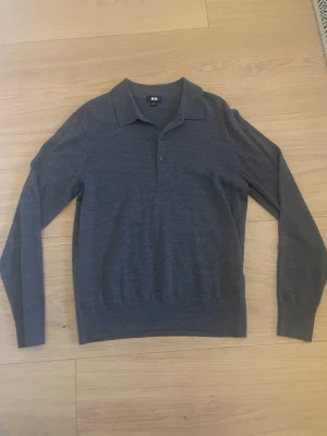 Grå långärmad pikétröja Uniqlo - Snygg grå långärmad pikétröja från Uniqlo i merinoull. Tröjan har klassisk krage, knäppning med tre knappar och ribbade muddar vid ärmslut och nederkant. Perfekt för en clean och stilren look.