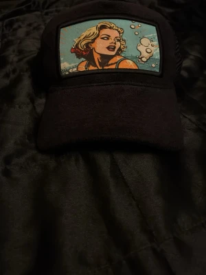 Svart keps med pop art-tryck - Snygg svart keps med ett färgstarkt pop art-tryck framtill som föreställer en blond person i serietidningsstil med blå bakgrund och bubblor. Kepsen har klassisk form och justerbar baksida. Perfekt för dig som vill sticka ut med en unik accessoar.