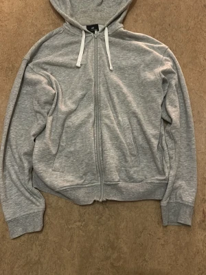 Grå zip hoodie från H&M Relaxed Fit - Snygg gråmelerad hoodie från H&M i relaxed fit. Tröjan har dragkedja framtill, två fickor, vit dragsko i huvan och ribbade muddar vid ärmar och nederkant. Perfekt för en chill och avslappnad stil. Inga defekter storlek S