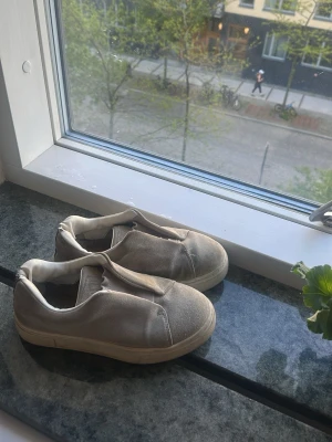 Beige slip-on sneakers utan snörning - Säljer ett par beige slip-on sneakers med rund tå och tjock vit sula. Skorna har en minimalistisk design utan snörning och är tillverkade i tygmaterial. Perfekta för en avslappnad och stilren look.