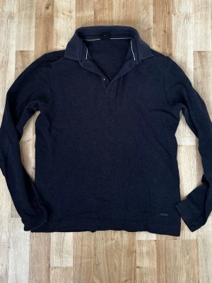 Svart långärmad tröja Hugo Boss - Snygg svart  långärmad tröja från Hugo Boss i slim fit-modell. Tröjan har klassisk krage med knappdetalj och är tillverkad i mjukt material. Perfekt för en stilren och avslappnad look.