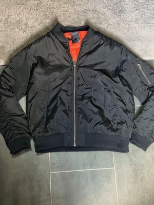 Svart bomberjacka med orange foder - Klassisk svart bomberjacka från Alpha Industries med orange foder. Jackan har ribbade muddar, dragkedja framtill, två snedställda fickor och en ärmficka med dragkedja. Tillverkad i glansigt nylonmaterial som ger en cool look.