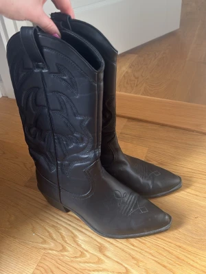 Svarta cowboy boots med brodyr - Snygga svarta cowboy boots med klassisk spetsig tå och dekorativ brodyr på skaftet och framtill. Liten skavank längst fram vid tån på den ena men det är verkligen inget som syns när man har på sig dem! 