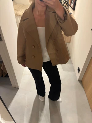 Kort beige dubbelknäppt kappa - Snygg kort kappa i beige brun färg med dubbelknäppning och bred krage. Kappan har en avslappnad passform och uppvikta ärmslut🌸