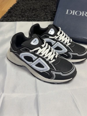 Svarta  sneakers med vita detaljer - Snygga svarta  sneakers med vita och ljusgrå detaljer samt stora CD-loggor på sidorna. Skorna har vita snören, svart mesh och syntetmaterial samt en vit och svart sula. Perfekta för dig som vill ha en trendig och exklusiv look.