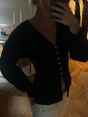 Svart kofta med guldknappar  - Stilren svart kofta med djup v-ringning och dekorativa knappar framtill. Figurnära passform och långa ärmar ger en snygg siluett. Perfekt att matcha med jeans eller kjol för en trendig look.