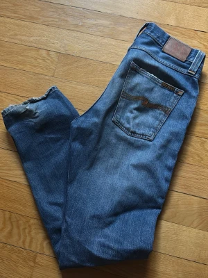 Nudie steady Eddie - Säljer ett par klassiska blå jeans från Nudie Jeans med snygga slitningar och den ikoniska vågade sömmen på bakfickan. Jeansen har normal passform och är gjorda i slitstarkt denim med läderpatch i midjan. 