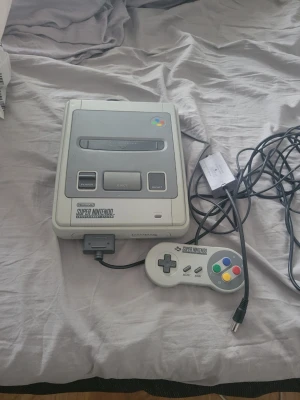 Super Nintendo SNES konsol med handkontroll - Klassisk Super Nintendo Entertainment System (SNES) konsol i grå plast med tillhörande original handkontroll. Konsolen har en rektangulär form och färgglada knappar på kontrollen. Perfekt för retrospel och nostalgi! Saknas ladd adapter. 