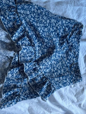 Blommig blå volangkjol - Supersöt kort kjol i blått med vitt blommönster och volangkant. Kjolen har omlott-look och är perfekt för sommarens alla äventyr. Lätt och luftig känsla, passar perfekt till en enkel topp eller stickad tröja.