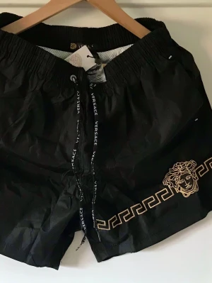 Svarta badbyxor från Versace - Snygga svarta badbyxor från Versace med guldfärgad Medusa-logga och klassiskt grekiskt mönster längs benet. Resårmidja med snörning där det står 'Versace' på banden. Perfekta för stranden eller poolen, riktigt lyxig känsla och stilren design.