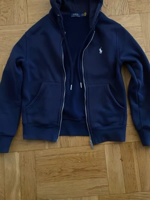 Mörkblå hoodie från Polo Ralph Lauren - Mörkblå hoodie med dragkedja från Polo Ralph Lauren. Klassisk design med vit broderad logga på bröstet, två fickor fram och justerbar huva med snören. Mjuk insida i fleece och ribbade muddar vid ärmar och nederkant. Perfekt för en avslappnad stil.