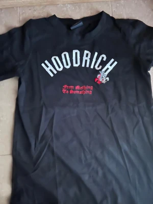 Svart HOODRICH t-shirt med tryck - Svart t-shirt från HOODRICH med stort vitt tryck fram och bak. På framsidan står det 'HOODRICH' och 'From Nothing To Something' i rött, samt en liten detalj med lilja. Baksidan har texten 'product of the streets' i stor stil. Klassisk passform och rund hals.