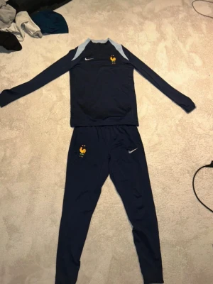 Frankrike Nike träningsset blå - Snyggt träningsset från Nike i mörkblått med ljusblå detaljer. Setet består av långärmad tröja och matchande byxor, båda med Frankrikes fotbollslandslagsmärke och Nike-logga. Perfekt för fotbollsträning eller chill. Materialet är troligtvis polyester för bästa komfort.