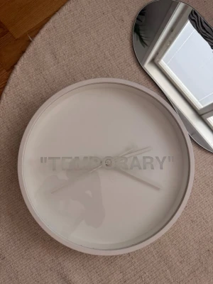 Off white klocka - Minimalistisk rund väggklocka i vit plast med texten 'TEMPORARY' tryckt på urtavlan. Klockan har enkla vita visare och saknar siffror, vilket ger en stilren och modern look. Perfekt som inredningsdetalj för dig som gillar unik design.