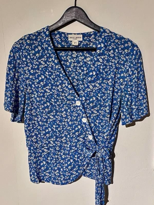 Kornblå blommig omlottblus med knytning 💙 - Säljer en supersöt kornblå omlottblus från Holly & Whyte by Lindex! Detaljerad med små vita blommor, fina knappar fram, långt knytband och stretchig resårrygg. Gjord i 100% skön och hållbar EcoVero-viskos. Skick: Nyskick! (Helt felfri). Mått: Armhåla-armhåla ca 45 cm, längd 56 cm, midja 40 cm. Se sista bilden!  Storlek: EUR S Köparen står för frakten. Samfraktar självklart! 📦✨