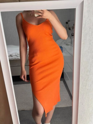 Ribbad orange klänning med slits - Säljer en ribbad, figurnära klänning i stark orange färg. Klänningen har smala axelband, v-ringning och en snygg slits framtill. Perfekt för dig som vill sticka ut med en färgstark look.