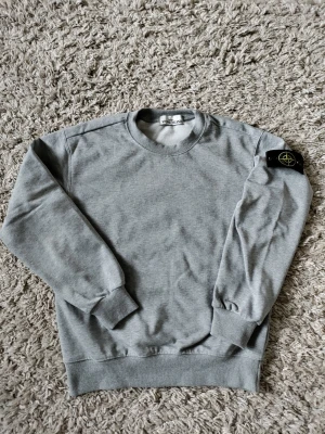Grå sweatshirt från Stone Island - Säljer en stilren grå sweatshirt från Stone Island med klassisk rund hals och ribbade muddar. Tröjan har lång ärm och den ikoniska logotypen på vänster ärm. Perfekt för dig som gillar streetwear och enkel design.