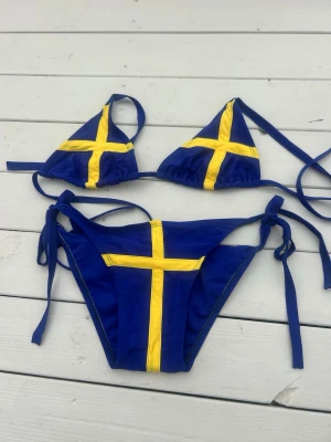 Blå bikini med svenska flaggan - Snygg blå bikini med gula kors som liknar svenska flaggan. Överdelen är trekantsmodell med knytband i nacken och ryggen, och underdelen har knyt i sidorna. Köpt utomlands, står ingen storlek men skulle tippa på S ungefär. Den har lite gröna ränder i de gula strecken som ni kan se på bilderna. 