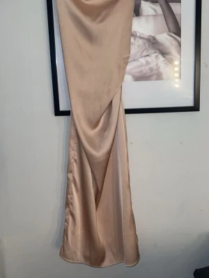 Champagnefärgad långklänning i satin - Elegant långklänning i champagnefärgad satin med omlott-detalj framtill. Klänningen har en mjuk glans och ett följsamt fall som ger en lyxig känsla. Perfekt för dig som vill ha en stilren och trendig look. Funkar även till balklänning. Djur finns i hemmet, tvättas innan leverans.🌼