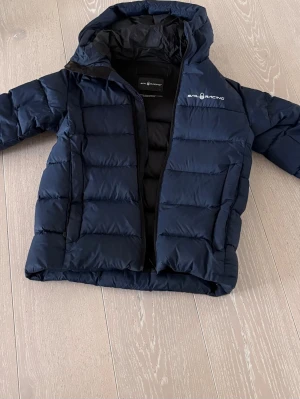 Mörkblå dunjacka Sail Racing - Snygg mörkblå dunjacka från Sail Racing med huva och dragkedja. Jackan har quiltad design och är fylld med 80/20 dun och fjäder, 600 fill power. Perfekt för kalla vinterdagar och har logga på bröstet.