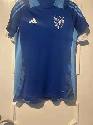 Blå IFK Norrköping träningstop - Snygg blå träningstop från IFK Norrköping med klubbmärke på bröstet och Adidas-logga. Tröjan har korta ärmar, rund hals och ljusblå paneler på sidorna samt axlarna. Tillverkad i ett lätt och ventilerande funktionsmaterial, perfekt för träning eller match.