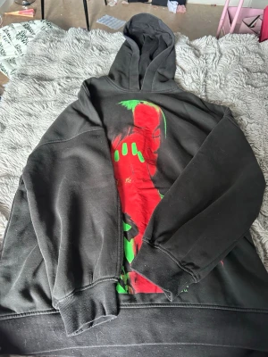Billie eilish hoodie - Säljer en svart hoodie med stor färgstark print på framsidan i rött och grönt. Motivet föreställer ett ansikte med neonfärgade detaljer och grafiskt mönster. Hoodien har huva och ribbade muddar vid ärmar och nederkant. Perfekt för dig som gillar streetwear och unika plagg.