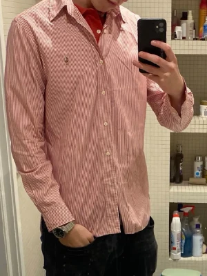 Rödvit randig skjorta från Ralph Lauren  - Snygg skjorta med röda och vita smala ränder, klassisk krage och knäppning framtill. Långärmad modell i bomull, perfekt för en stilren look. Liten broderad logga på bröstet ger extra detalj.