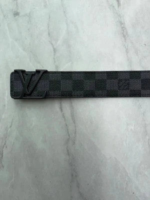 Louis Vuitton rutigt bälte svart/grå - Säljer ett Louis Vuitton bälte med klassiskt rutigt mönster i svart och grått. Bältet har en stor LV-logga som spänne och är gjort i skinn. Perfekt accessoar för att lyfta din outfit.