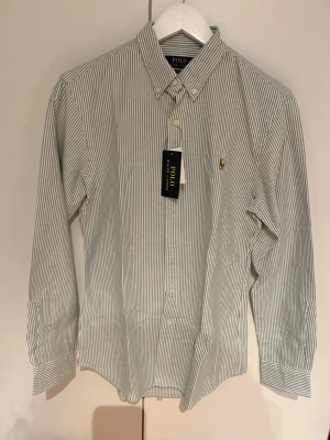 Randig skjorta från Polo Ralph Lauren - Snygg vit skjorta med tunna gröna ränder från Polo Ralph Lauren. Slim fit-modell med klassisk krage, knappar framtill och broderad logga på bröstet. Tillverkad i mjuk bomull och har långa ärmar med knapp vid manschetten.
