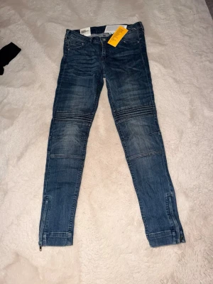Blå bikerjeans med dragkedjor - Säljer ett par blå bikerjeans från H&M med coola sömdetaljer över knäna och dragkedjor vid bensluten. Jeansen har en smal passform och klassisk femficksdesign. Perfekta för en trendig och avslappnad stil.