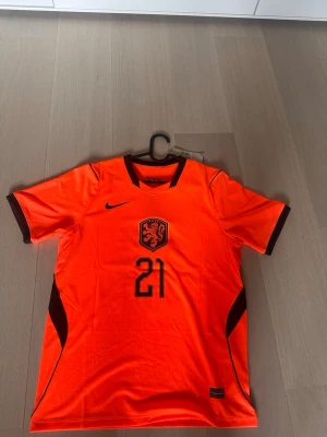 Holland matchtröja Nike nr 21 De Jong - Snygg orange Holland matchtröja från Nike med nummer 21 och De Jong på ryggen. Tröjan har svarta detaljer, korta ärmar och är tillverkad i ett lätt, ventilerande funktionsmaterial. Perfekt för fotbollsträning eller att bära som supporter.