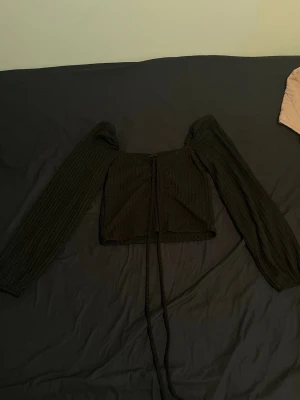 Svart ribbad croppad topp med snörning - Trendig svart croppad topp med långa ärmar och snygg snörning framtill. Toppen är ribbad och har en figurnära passform som passar perfekt till både jeans och kjol. Perfekt för dig som vill ha en stilren men ändå edgy look.