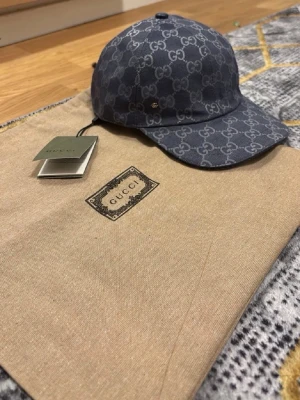 Blå GG-monogram keps från Gucci - Snygg blå keps från Gucci med det klassiska GG-monogrammet över hela kepsen. Kepsen har böjd skärm och justerbar rem bak. Tillverkad i canvasmaterial och levereras med originalpåse och taggar. Perfekt för dig som gillar exklusiva accessoarer.