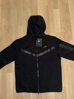 Svart Nike Tech Fleece hoodie - Svart Nike Tech Fleece hoodie med dragkedja och huva. Snygga svarta detaljer i form av diagonala band över bröstet och ärmen. Klassisk Nike-logga på bröstet. Perfekt för en sportig och stilren look.