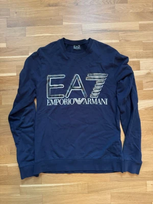Mörkblå EA7 Emporio Armani sweatshirt - Snygg mörkblå sweatshirt från EA7 Emporio Armani med stort vitt logotryck på bröstet. Tröjan har rund halsringning, långa ärmar och ribbade muddar vid ärmslut och nederkant. Perfekt för en stilren och sportig look.