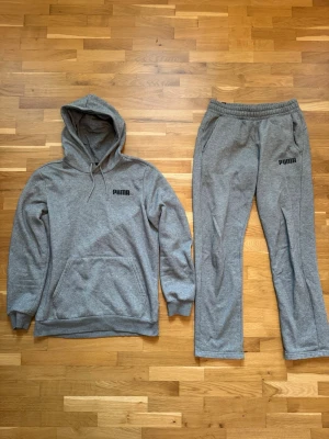 Grått set från Puma med hoodie och byxor - Säljer ett grått mjukisset från Puma med matchande hoodie och byxor. Hoodien har luva, magficka och ribbade muddar. Byxorna har resår i midjan och sidofickor. Båda delarna har Puma-logga tryckt framtill. Perfekt för en avslappnad look.
