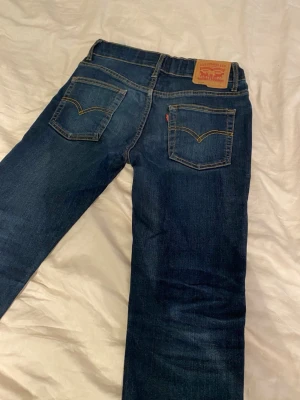 Mörkblå Levi's jeans med raka ben - Säljer ett par klassiska mörkblå jeans från Levi's med raka ben och femficksmodell. Jeansen har kontrastsömmar, läderpatch bak i midjan och den ikoniska röda Levi's-taggen på bakfickan. Perfekta för en avslappnad och tidlös stil.