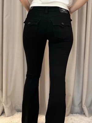 Svarta bootcut jeans True Religion - Säljer ett par svarta bootcut jeans från True Religion. Jeansen har klassiska bakfickor med lock och knapp, samt en snygg passform som sitter tight upptill och släpper ut vid benen. 