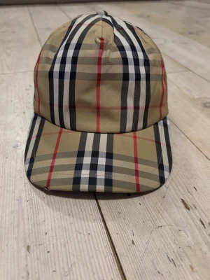 Beige rutmönstrad keps - Snygg keps i klassiskt beige rutmönster med svarta, vita och röda linjer. Kepsen har böjd skärm och justerbar baksida. Tillverkad i ett lätt tygmaterial som ger en stilren och trendig look.