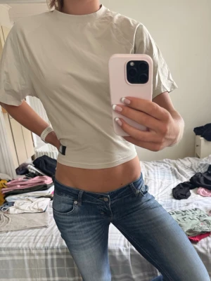 Beige cropped t-shirt - Säljer en beige cropped t-shirt med tight  passform över midjan och korta ärmar. T-shirten har en hög halsringning och en liten svart detalj nertill på sidan. Perfekt till jeans eller kjol för en clean och enkel look.