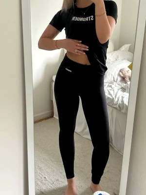 Svarta leggings från Stronger - Säljer ett par svarta leggings från Stronger med hög midja och tight passform. De är tillverkade i ett stretchigt material som sitter skönt och följer kroppen. Perfekta för träning eller till vardags. Diskret logga framtill vid midjan.